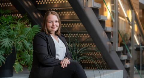 Professionally dressed student sits with pursed lips on a granite bench. In the background, a large, green plant, a staircase & soft lighting from wall sconces.