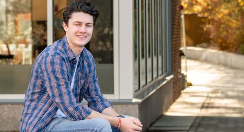Analytical economics and student-designed major Sam Croteau sits outside the UNH Paul College building on the edge of a flowerbed, smiling at the camera.