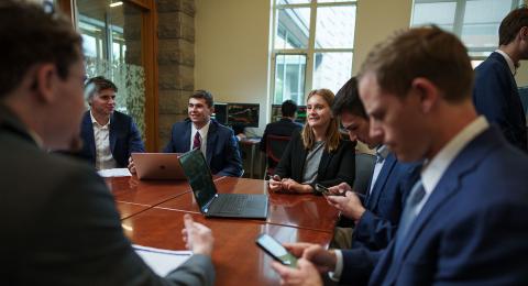 Professionally dressed students sit around a large table. Some hold phones & some have  laptops, as they work collaboratively. 