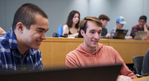 Two male students sit at a desk with laptops opening, smiling. In the blurred background, four students sit at a desk with their laptops.