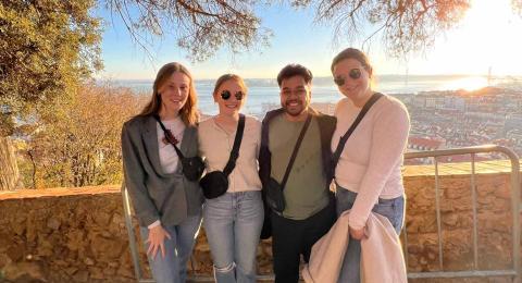 Four students pose in front of a stone wall, smiling. In the background is bright sunlight, city roofs, and the ocean in the distance. Trees hang overhead.