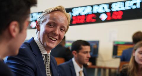 Professionally dressed students working in a classroom. In the center, a blonde, male student smiles at his classmate. In the background, a stock market ticker runs data on a screen.