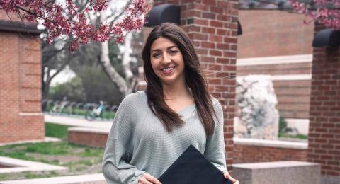 UNH Economics major Emily Bolognino stands on UNH campus in front of a blossoming tree, smiling at the camera, holding a graduation cap.