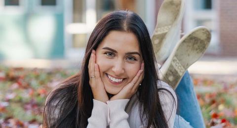UNH International Business and Economics major and transfer student Gabriella Caterino lies on a blanket on top of an autumn-leaf strewn lawn, hand under her chin and legs kicked up behind her in the air, grinning at the camera.