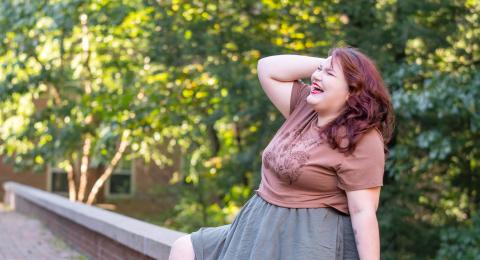 Student Laura Earle sits on a stone wall, hand in her hair, laughing. Her feet rest on a stone walkway and trees and dappling sunlight are in the background. 