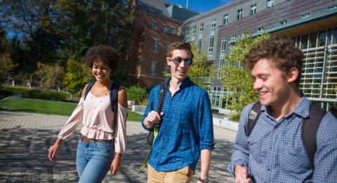 Three students stroll in the stone-paved courtyard, smiling with backpacks on. In the background, is grass and the exterior of the Paul College building.