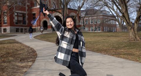 Hannah Wirth raises her graduation cap up in celebration in front of UNH's Thompson Hall.