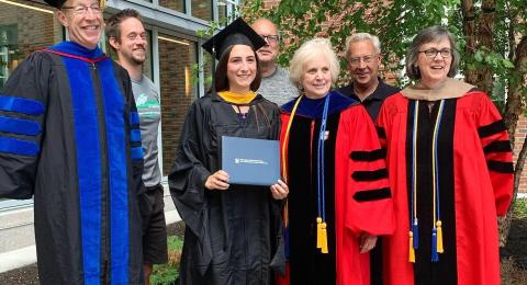 Faculty in graduation robes pose with a student in her cap and gown as she holds her diploma. There is a tree and Paul College in the background.