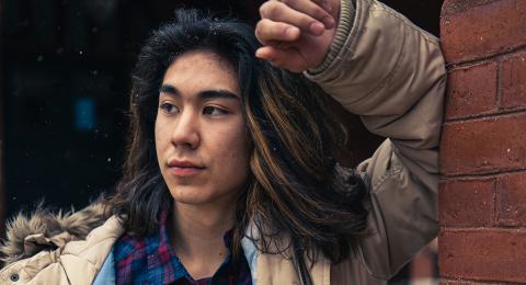 UNH Paul College student Kiyoshi Dustin leans on a brick wall outside Thompson Hall. He is wearing a winter coat with a fur lined hood, and a plaid button down.