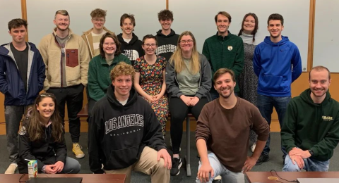 Students on the UNH Paul College Artifex club pose in a classroom with desks in the foreground and whiteboard in the background.