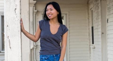 Student smiles, with her head turned to the left, as she places a hand on a column on a front porch. She has long, dark hair, and is dressed casually.
