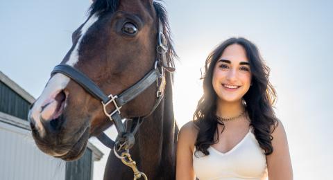 UNH Paul College student Sam Bhacka stands alongside her UNH horse and smiles. The space in between her and her horse is haloed by bright sunlight.