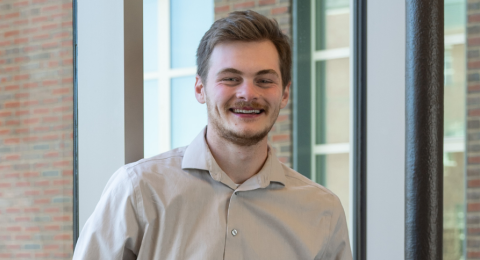 Male student smiles wearing a tan dress shirt. He has brown hair and a beard. Behind him is windows, and beyond, a brick building. 