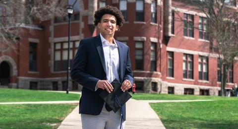 Tejun Celestin stands outside Thompson Hall with a graduation cap.