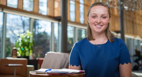 Smiling student Devin McMahon sits with a small desk with a notebook and pen in the foreground. Stippling light streams through windows in the building's atrium.