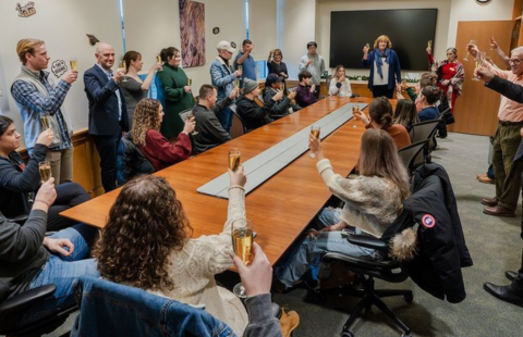 Students sit around a long conference table with a glass of champagne raised, with Dean Lucy Gilson at the head of the table also raising a glass.