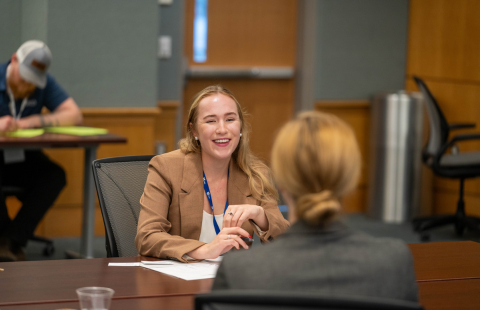A smiling female student in professional attire interviews with a business woman. They are seated face to face in a classroom.