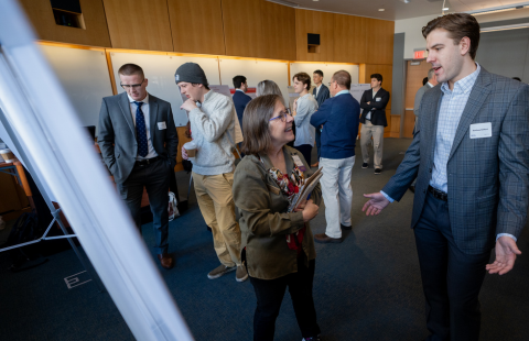 Student engages with professor in front of his research poster in a classroom surrounding by other students presenting at the URC