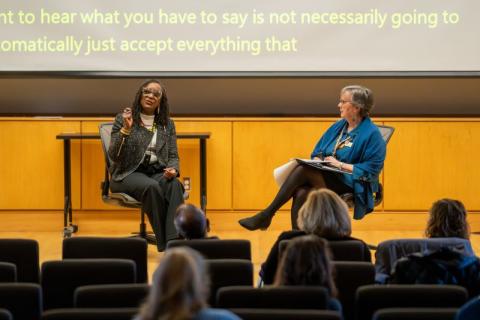 Two woman sit on stage presenting to a group in an auditorium.