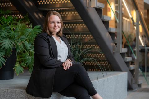 Professionally dressed student sits with pursed lips on a granite bench. In the background, a large, green plant, a staircase & soft lighting from wall sconces.