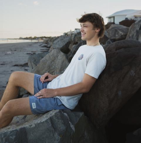 UNH Marketing and Finance Major Gavin Cieplik leans against some rocks on a local beach, smiling at something off camera.
