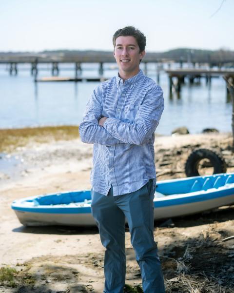 Paul College student stands with arms crossed, smiling. He wears a stripped button down & dress pants. In the background, a boat on land, and piers in the water.