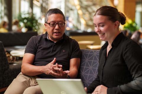 An older man sits and talks with a student in UNH Paul College's Great Hall, gesturing in a way that suggests he's giving advice on what's on her laptop.