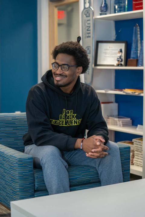 UNH Economics major Christian Katumba sits in a chair at the UNH ECenter, smiling at something off camera. He has an afro pick in his hair and is wearing a sweatshirt that says, "I am Black excellence."