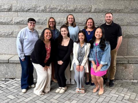 Group of nine people posing together for a photo in front of a gray stone wall, dressed in business casual attire