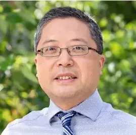 Professional headshot of Jun Li wearing glasses, light blue shirt, and striped tie, smiling against green foliage background.