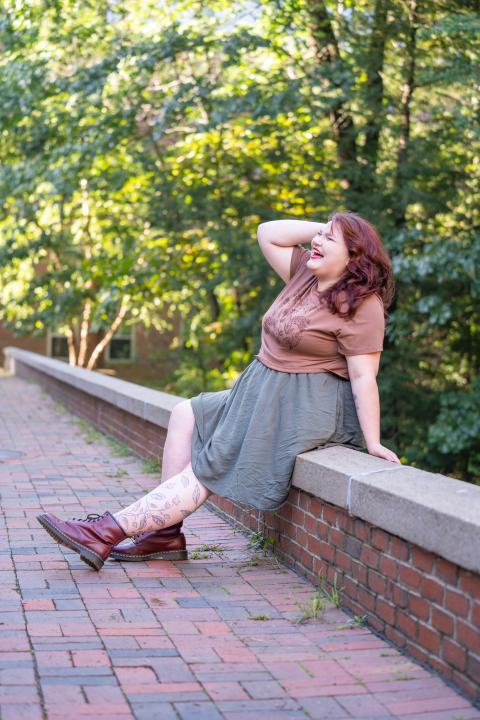 Student Laura Earle sits on a stone wall, hand in her hair, laughing. Her feet rest on a stone walkway and trees and dappling sunlight are in the background.