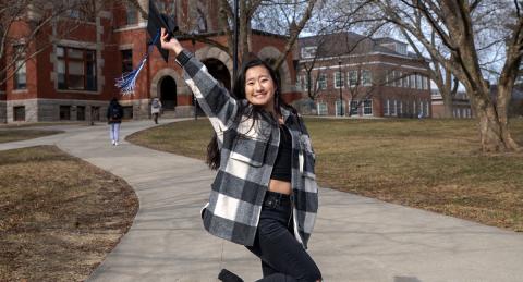 Hannah Wirth raises her graduation cap up in celebration in front of UNH's Thompson Hall.