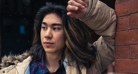 UNH Paul College student Kiyoshi Dustin leans on a brick wall outside Thompson Hall. He is wearing a winter coat with a fur lined hood, and a plaid button down.