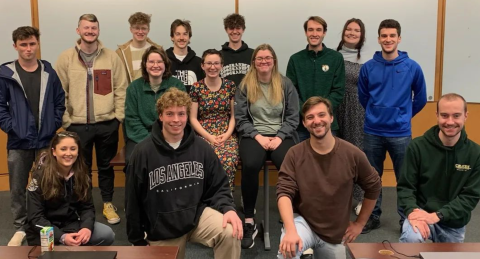 Students on the UNH Paul College Artifex club pose in a classroom with desks in the foreground and whiteboard in the background.