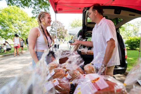 Briella Hirsch chats with a vendor at the Exeter farmer's market. A female student stands at a table of bread at an outdoor farmer's market talking to an older man.