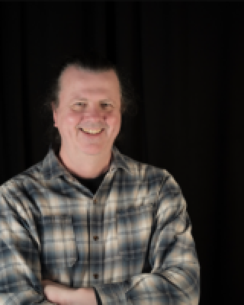 Headshot of Paul Harvey, a man with a plaid shirt, arms folded, dark hair, and a black background