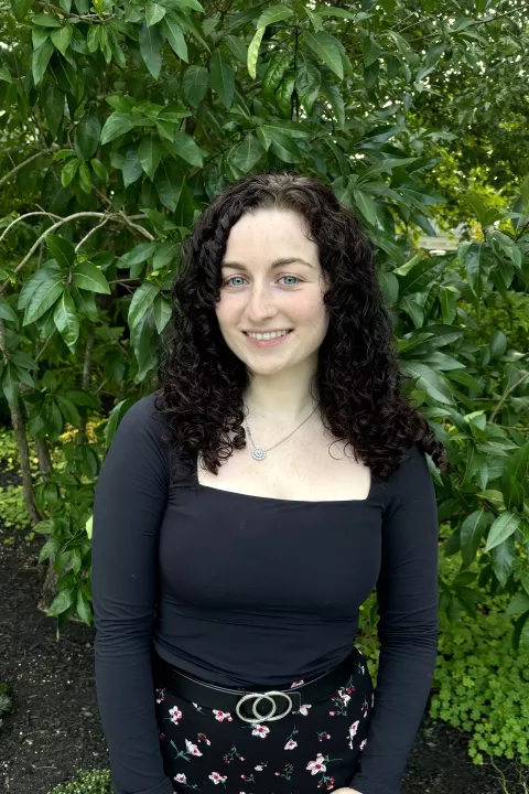 Photo of Sarah DeSimone standing outside. She has dark, shoulder length brown hair and in the background is a green, leafy tree.