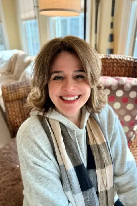 Photo of Noelle Lee, a woman wearing a gray hoodie and a scarf sitting on a upholstered chair with windows in the background