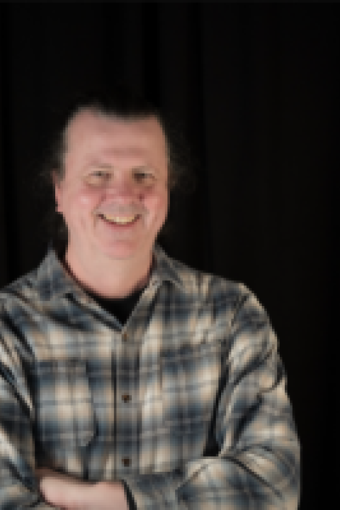 Headshot of Paul Harvey, a man with a plaid shirt, arms folded, dark hair, and a black background