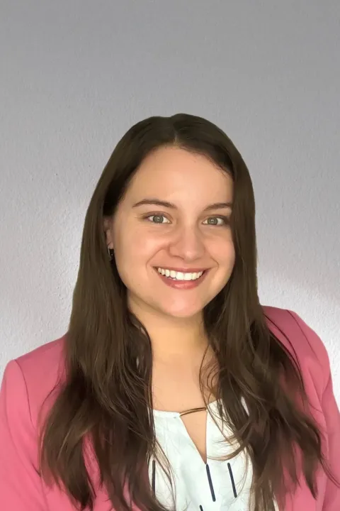 Headshot of Peyton Rockcliffe, a woman with long brown hair wearing a white top and pink blazer, with a gray background