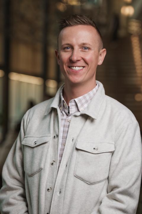 Headshot of Jon Savey. White male wearing a plaid button down with gray button up layer on top. With short from hair. In the background,  a blurred room.